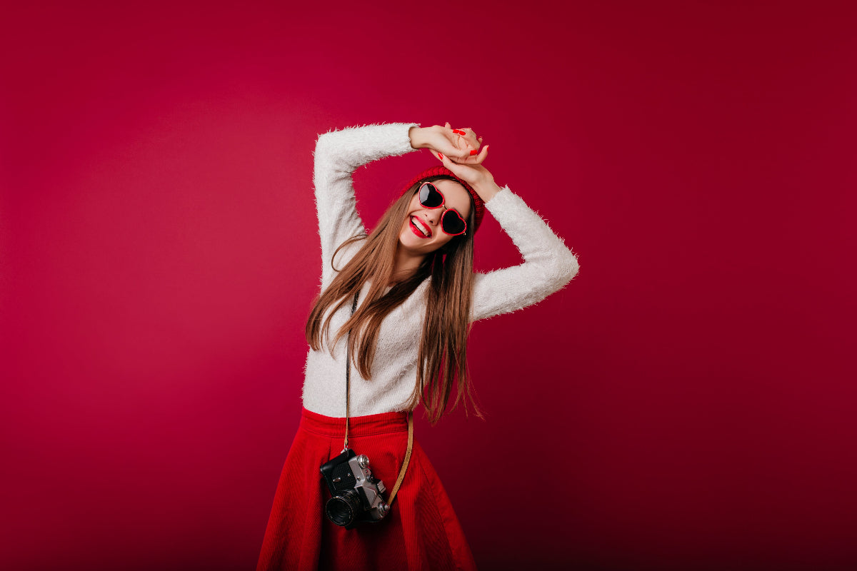 Woman in a white sweater and red skirt posing with a camera against a pink background
