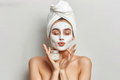 Woman applying facial mask with a towel on her head against a plain background