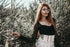 Woman standing among tall plants wearing sunglasses and a black top with a white skirt.